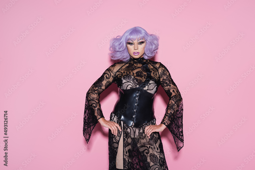Fototapeta premium eccentric drag queen in purple wig and black lace dress looking at camera on pink.