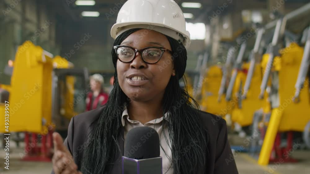 African American female journalist in hardhat and formalwear standing in heavy machinery factory, looking at camera and speaking in microphone during live TV stream African American female journalist in hardhat and formalwear standing in heavy machinery factory, looking at camera and speaking in microphone during live TV stream