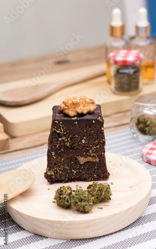Chocolate cupcake muffins with cannabis buds in wooden spoon, oil pot and cooking jars. Marijuana hemp in food dessert. Chocolate herb muffins cooking. Marijuana cake on a brown wooden table.