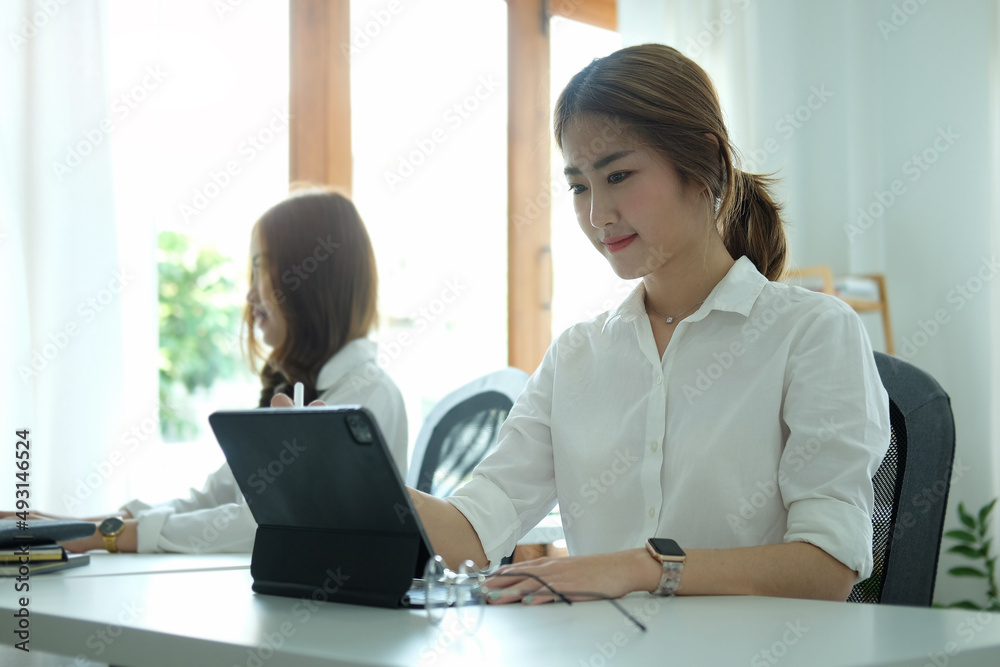 Charming asian woman office worker using computer laptop while sitting with her colleague in office. Charming asian woman office worker using computer laptop while sitting with her colleague in office.