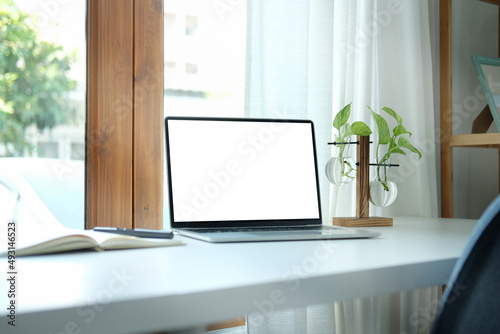 Computer laptop with empty screen  book and houseplant on white table in bright living room.