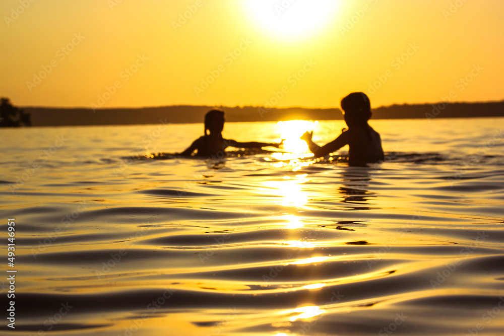 Children frolicking playing in water of river, lake, sea at sunset. Preschoolers on summer vacation. Silhouettes of kids have joy on setting yellow sun background. Soft small waves rolling onto shore.