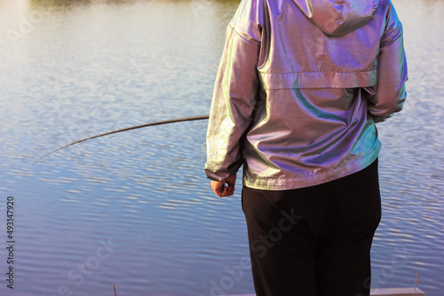 Fisherwoman with a yellow fishing rod by a pond, river, lake on a sunny spring day. Woman back view catching fish. Active leisure and recreation in nature reserve. Angling on a bank. Blue water.