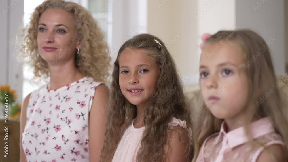 Portrait of pretty charming girl smiling looking at camera sitting indoors with sibling and woman. Happy confident Caucasian kid posing in slow motion with blurred mother and sister Portrait of pretty charming girl smiling looking at camera sitting indoors with sibling and woman. Happy confident Caucasian kid posing in slow motion with blurred mother and sister