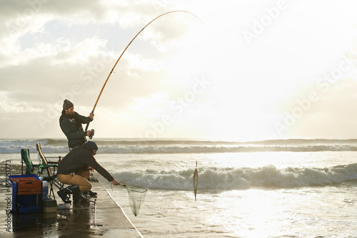 Ive got the big one. Shot of two young men fishing off a pier.