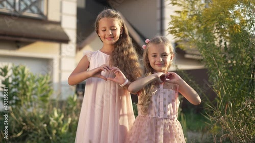 Medium shot of Caucasian smiling sisters gesturing heart shape with hands looking at camera. Portrait of charming girls posing at sunset on front of house outdoors on spring summer day photo