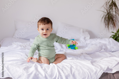 baby in a green cotton bodysuit is sitting on a white bed with a colored toy car. child is playing