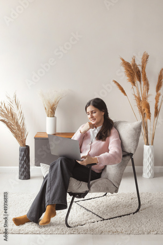 Attractive confident woman 30 years old working remote on laptop from cozy home sitting in comfortable chair with trendy interior