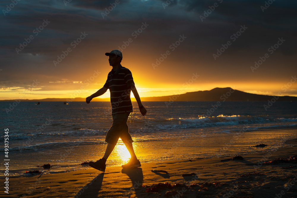 Silhouette man walking on Milford beach at sunrise, Rangitoto Island in the background, Auckland.