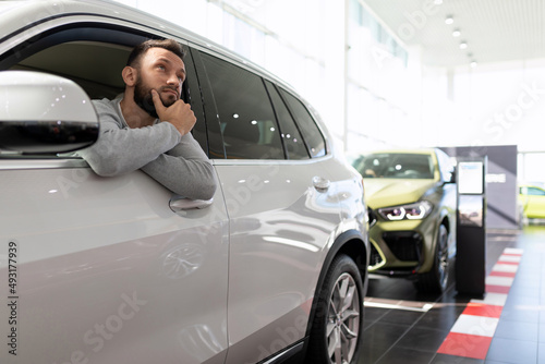 a buyer in a car dealership sits in an SUV and thinks about buying and insuring a new car © Ivan Traimak