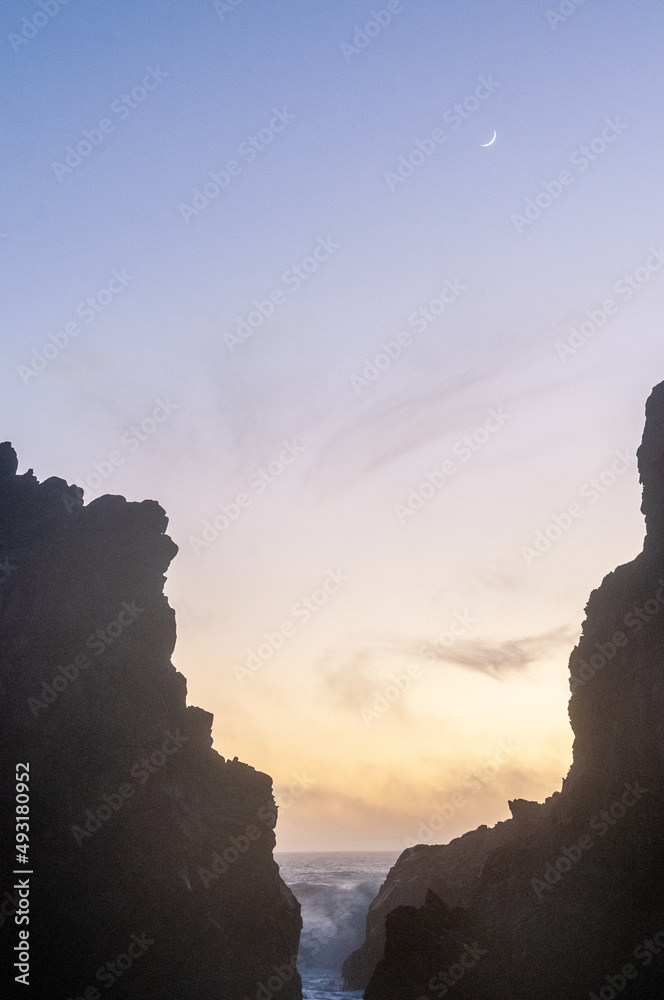 Incoming waves from the pacific ocean are breaking through the rocks at pfeiffer beach, around sunset. A small crescent moon illuminates the sky.