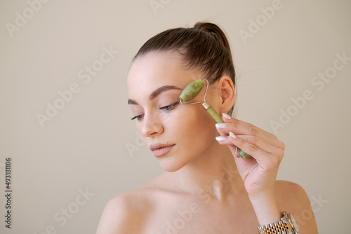 Close-up portrait caucasian pretty woman doing elevator lift smoothing anti-aging massage with green stone roller, isolated over beige background with copy space. Facial Skin Care Concept photo