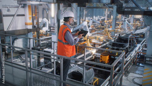 High angle of mature male engineer in uniform and hardhat using tablet to control robotic arms during work on contemporary car factory photo