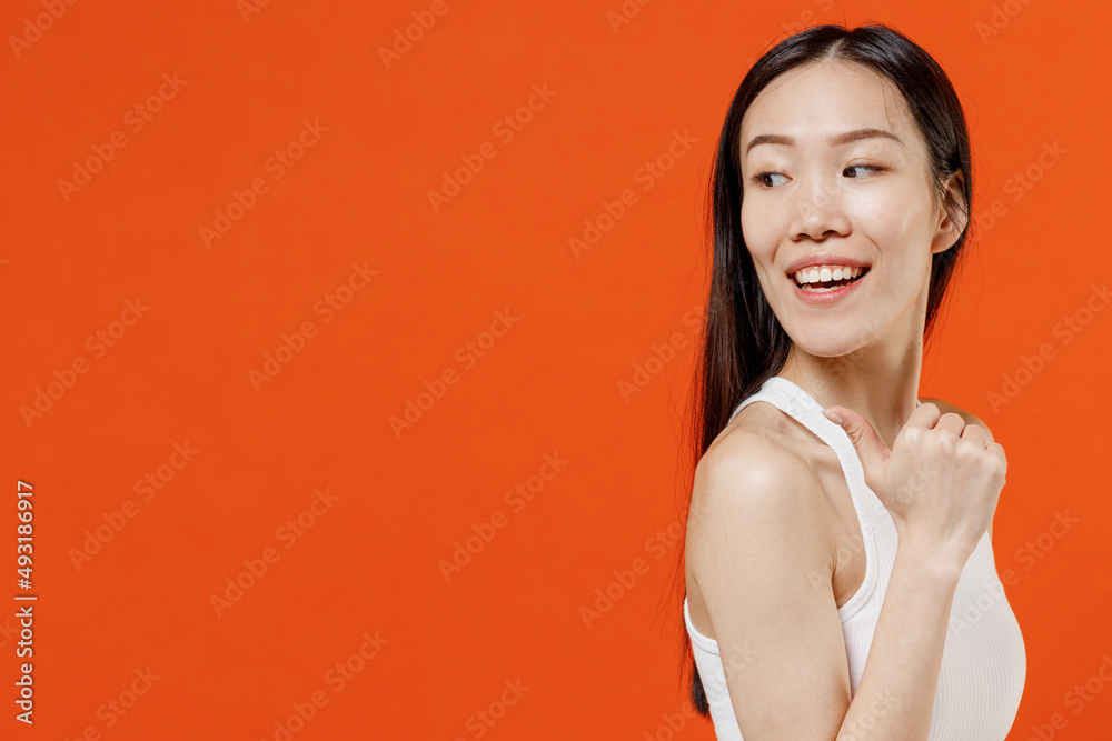 Joyful fun exultant young woman of Asian ethnicity 20s year old in white tank top pointing thumb finger behind on workspace area copy space mock up isolated on plain orange background studio portrait