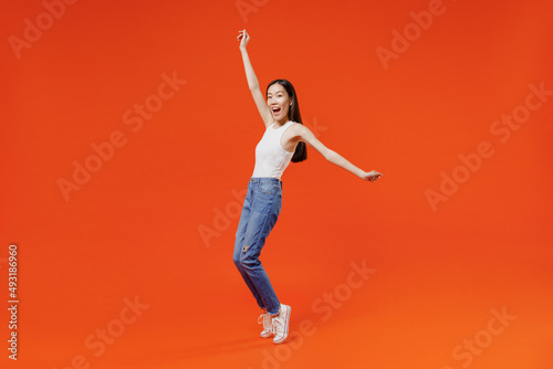 Full size body length fun young woman of Asian ethnicity 20s year old in white tank top standing on toes dancing lean back have fun spreading hands isolated on plain orange background studio portrait