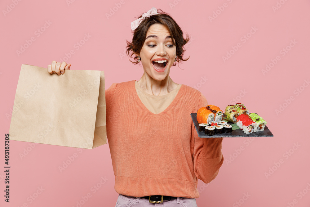 Young surprised woman in sweater hold makizushi sushi roll served on black plate traditional japanese food brown clear blank craft paper takeaway bag mock up isolated on plain pastel pink background Young surprised woman in sweater hold makizushi sushi roll served on black plate traditional japanese food brown clear blank craft paper takeaway bag mock up isolated on plain pastel pink background