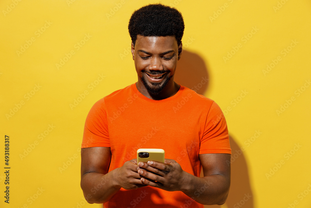 Young smiling happy cheerful man of African American ethnicity 20s wear orange t-shirt hold in hand use mobile cell phone isolated on plain yellow background studio portrait. People lifestyle concept. Young smiling happy cheerful man of African American ethnicity 20s wear orange t-shirt hold in hand use mobile cell phone isolated on plain yellow background studio portrait. People lifestyle concept.