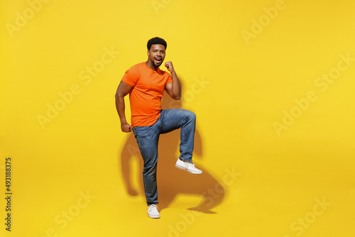 Full body happy young man of African American ethnicity 20s wear orange t-shirt doing winner gesture celebrate clenching fists say yes raise up leg isolated on plain yellow background studio portrait
