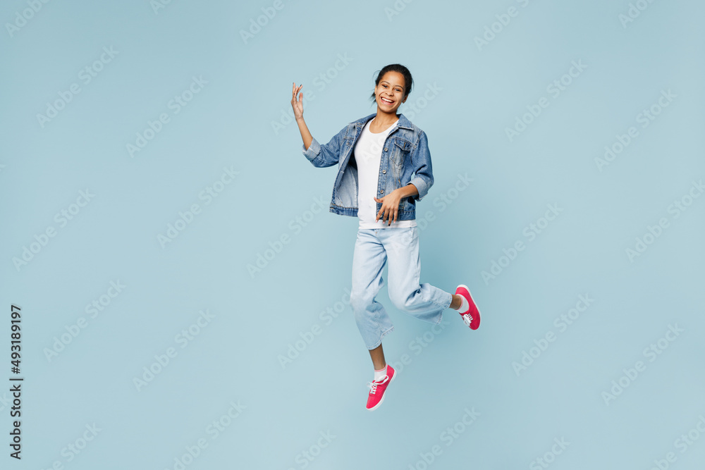 Full body happy little kid teen girl of African American ethnicity 12-13 years old in denim jacket play guitar jump high look camera isolated on pastel plain light blue background. Childhood concept.