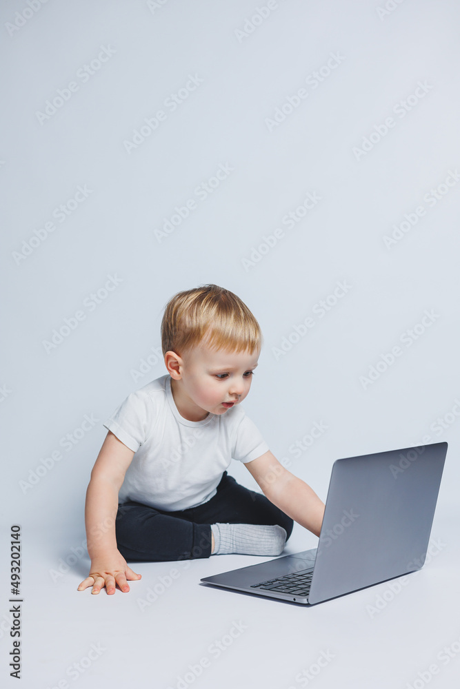 A little boy 3-4 years old sits with a laptop on a white background. A child in a white T-shirt and black trousers looks at a laptop. Modern children A little boy 3-4 years old sits with a laptop on a white background. A child in a white T-shirt and black trousers looks at a laptop. Modern children