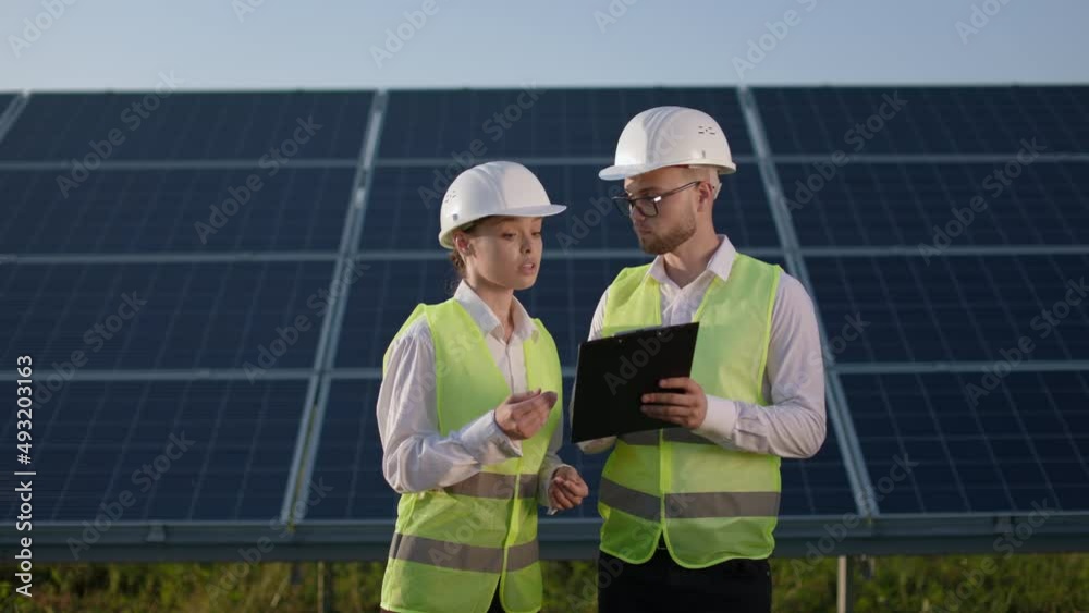 Caucasian man and woman in white helmet and safety vest, standing together on solar farm, talking and looking on clipboard. Concept of cooperation, green energy and ecology. Caucasian man and woman in white helmet and safety vest, standing together on solar farm, talking and looking on clipboard. Concept of cooperation, green energy and ecology.