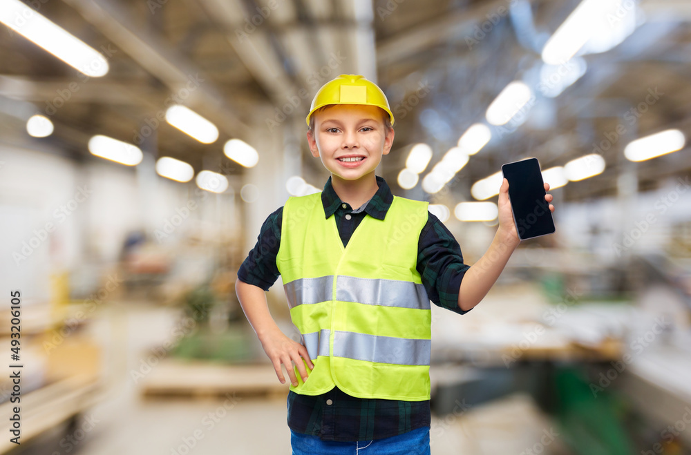 industry, manufacture and profession concept - happy smiling little boy in protective helmet and safety vest with smartphone over workshop background industry, manufacture and profession concept - happy smiling little boy in protective helmet and safety vest with smartphone over workshop background