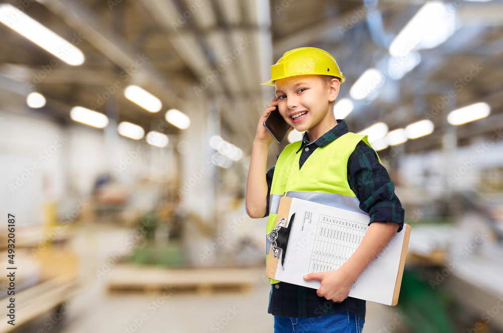 industry, manufacture and profession concept - happy smiling little boy in protective helmet and safety vest with clipboard calling on smartphone over workshop background