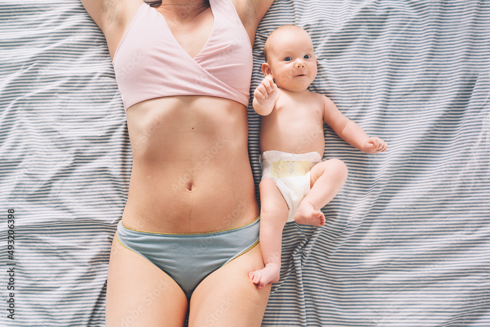 The postpartum (or postnatal) period. Young mother with visible postpartum  body marks and her cute little baby. Two months baby lying near at mother  belly on bed. Skin to skin. Photos
