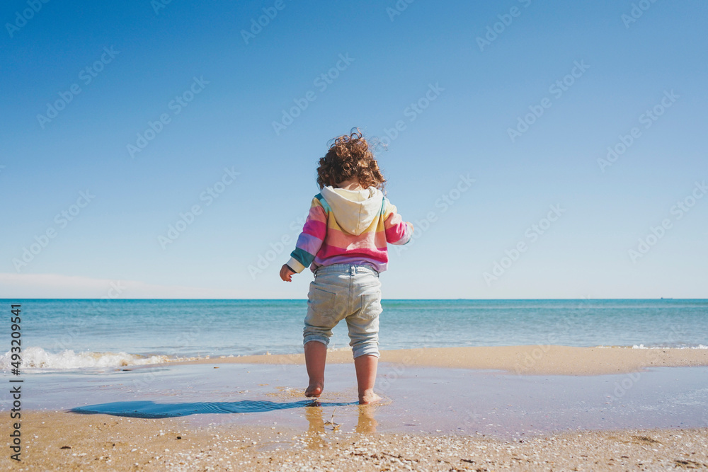 Wide view of a little girl wearing rainbow hoodie playing at the beach