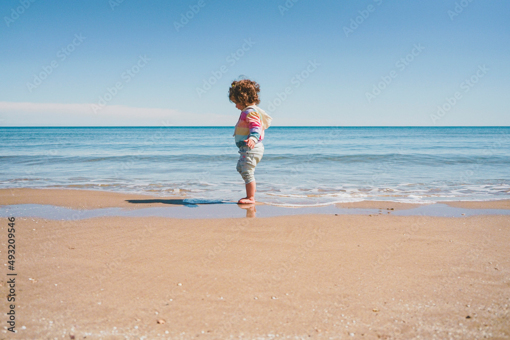 Wide view of a little girl wearing rainbow hoodie playing at the beach