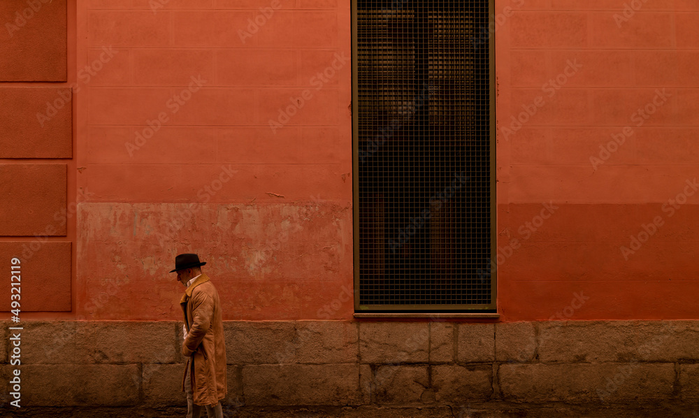 Adult man in hat and coat walking in front of red wall on street during sand storm day. Madrid, Spain Adult man in hat and coat walking in front of red wall on street during sand storm day. Madrid, Spain