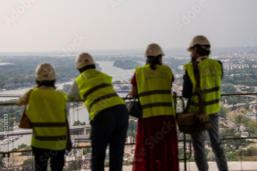 Diverse team of specialists with safety equipment vest and helmets observe work progress on construction site and enjoy beautiful city view from high-rise building, skyscraper.