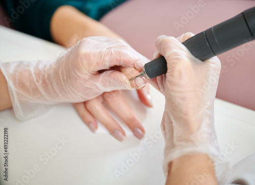 Manicurist using cuticle remover device while doing hardware manicure in beauty salon. Close up of female manicure master in sterile gloves removing cuticle on woman hand with electric nail drill.