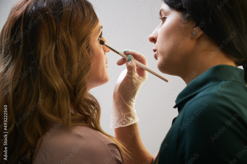 Close up of female beauty specialist using cosmetic brush while applying foundation or concealer under woman eye. Makeup artist doing professional makeup for client in visage studio.