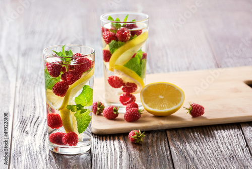Detox fruits infuse water with ice, raspberries, lemon, mint on wooden background. Refreshing summer cocktail, selective focus.