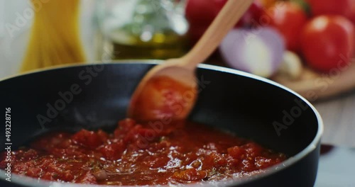 Close up of woman hand stirs with spoon boiling hot tomato sauce, cooked on stove. Fresh vegetables background. photo