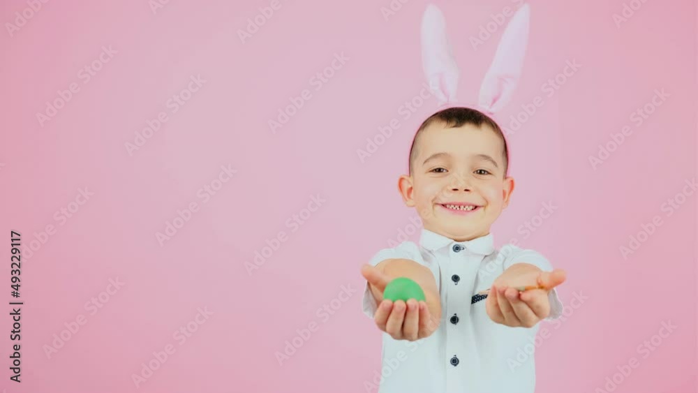 One little adorable boy with bunny ears on head showing for the camera green egg and brush he holding in his hands on pink background and copy space. One little adorable boy with bunny ears on head showing for the camera green egg and brush he holding in his hands on pink background and copy space.