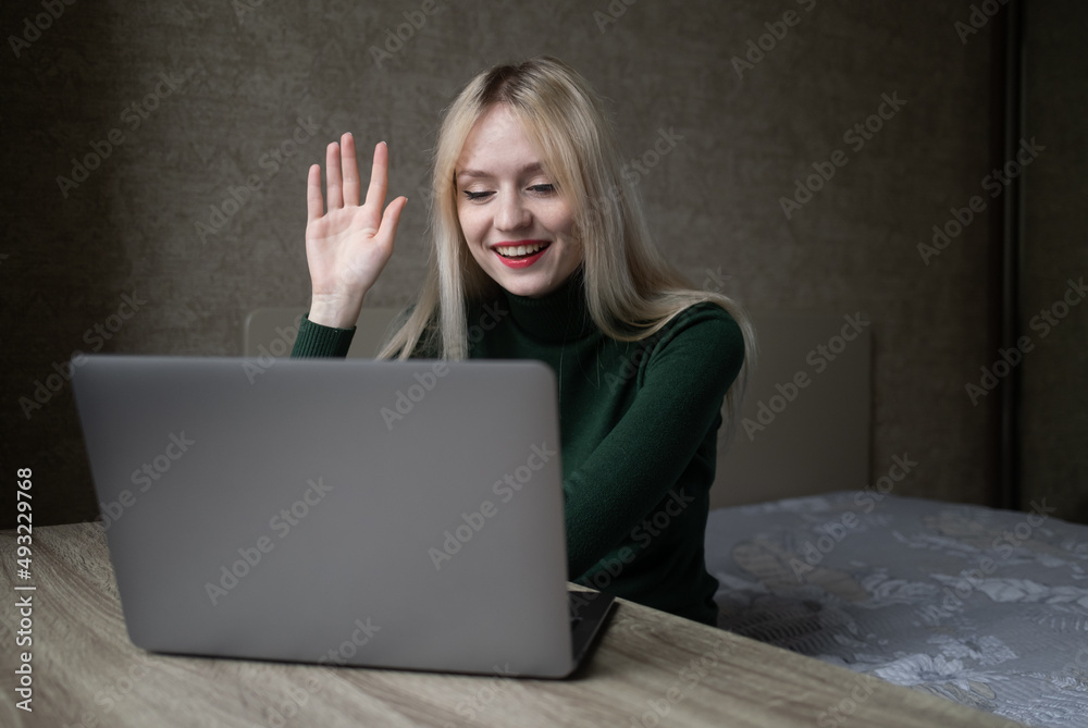 Emotional cheerful blonde girl in green golf is talking via video link at an online conference. Works at home with a laptop. Sits at the table in a positive mood in the room. Online learning. High Emotional cheerful blonde girl in green golf is talking via video link at an online conference. Works at home with a laptop. Sits at the table in a positive mood in the room. Online learning. High