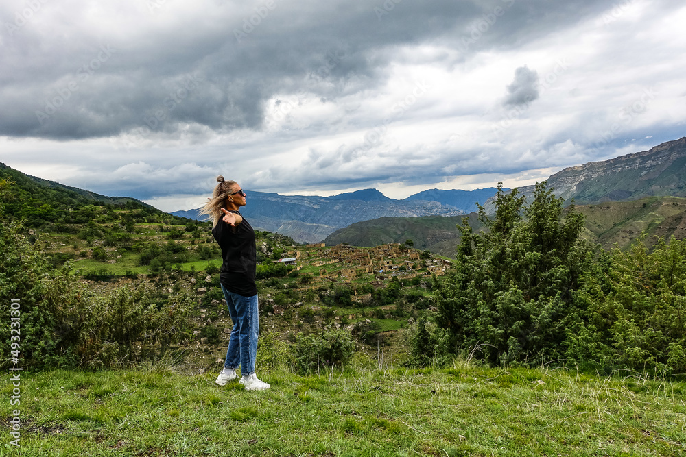 A girl on the background of the village of Kurib in the Caucasus mountains, on top of a cliff. Dagestan Russia June 2021