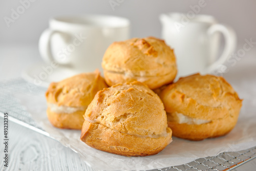 Homemade profiteroles on a metal grid and white paper on a white wooden background. Behind are a teacup and a milk jug.
