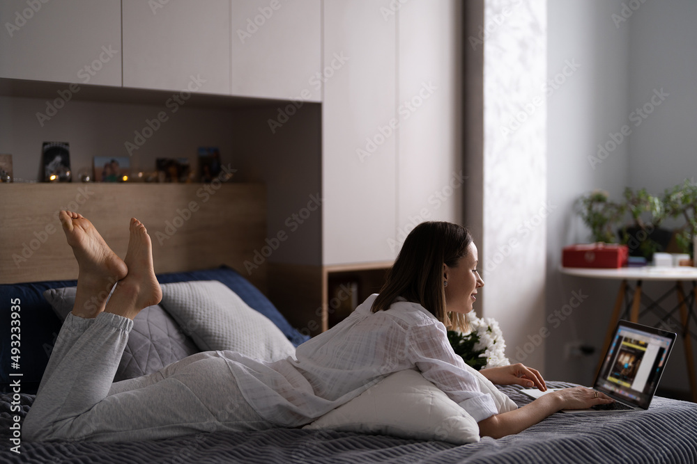 Young woman sitting on the bed wearing pajamas, with pleasure enjoying white flowers, chatting using laptop Young woman sitting on the bed wearing pajamas, with pleasure enjoying white flowers, chatting using laptop