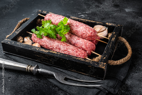 Fresh raw beef meat kebabs sausages on skewers in wooden tray. Black background. Top view