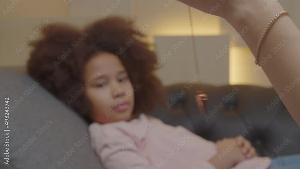 Close-up of female hand of psychologist hypnotizing lovely little black girl using pendulum, to create state of focused attention and increased suggestibility in treatment of psychological disorder.