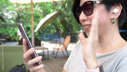 Stylish short hair happy Asian woman videocall, smile, chat with smartphone and wave hand to friends and family at beautiful outdoor cafe with trees in background. Social distancing and social media.