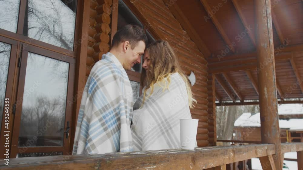 A guy and a girl wrapped in a blanket, standing at a wooden house in winter. Romance