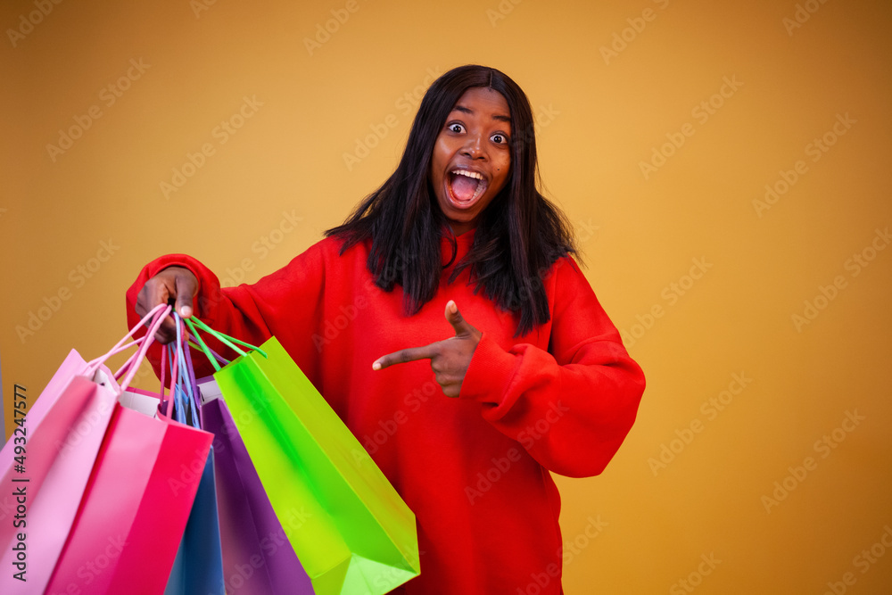 Smiling African-American woman in a red hoodie pointing her finger at colorful shopping bags and looking at the camera on a yellow background with copy space