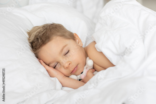 Beautiful blond toddler child, boy, sleeping in bed with teddy bear toy