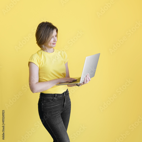 Young caucasian woman in yellow t-shirt and black jeans with laptop one bright yellow background. Business woman or college student concept.