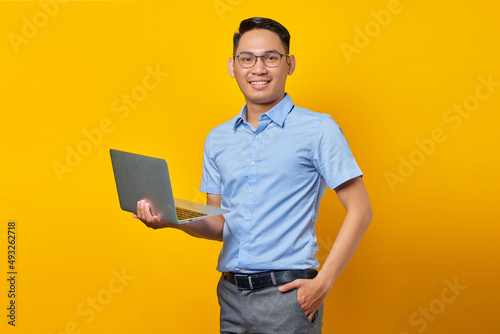 Portrait of smiling handsome Asian man in glasses holding laptop and looking at camera isolated on yellow background. businessman and entrepreneur concept
