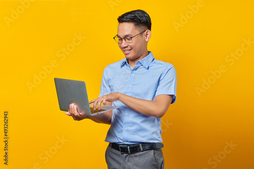 Portrait of cheerful handsome Asian man in glasses using laptop isolated on yellow background. businessman and entrepreneur concept